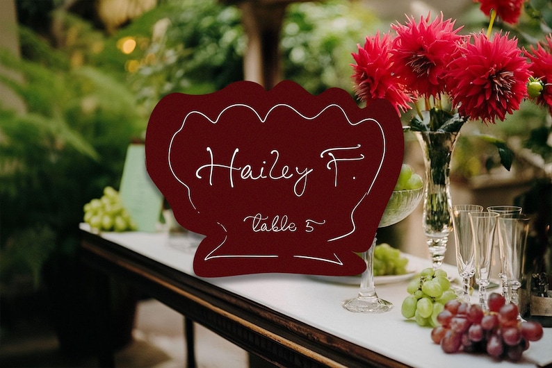 May include: A burgundy-colored place card with the name "Hailey F." and "table 5" in white script. The card has a decorative scalloped edge. The background includes red flowers, grapes, and glassware, suggesting a wedding or event setting.