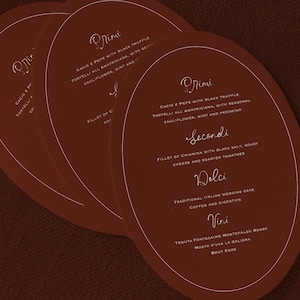 May include: Three oval-shaped menu cards in a warm brown hue, stacked slightly to reveal the text. The cards feature elegant, handwritten-style text in a light gold color, with menu items like "Cacio e Pepe" and "Fillet of Chianina".