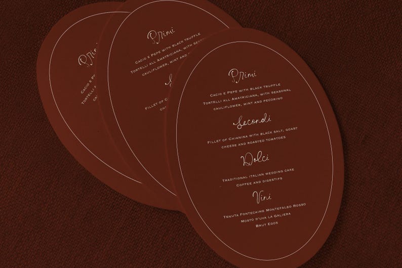 May include: Three oval-shaped menu cards in a warm brown hue, stacked slightly to reveal the text. The cards feature elegant, handwritten-style text in a light gold color, with menu items like "Cacio e Pepe" and "Fillet of Chianina".