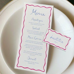 Puede incluir: Una tarjeta de menú blanca con un borde rosa, que presenta un menú escrito a mano para aperitivos, ensalada, plato principal y postre. El menú incluye platos como "Sonnichetta con glaseado balsámico" y "Pastel de boda de tiramisú italiano tradicional". Una tarjeta blanca separada con un borde rosa dice "Lactitia Mesa 1".