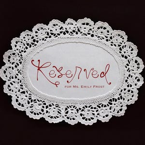 May include: White, oval-shaped doily with a delicate lace pattern. The center features the word "Reserved" in red script, followed by "for Ms. Emily Frost". The doily is set against a dark brown background.
