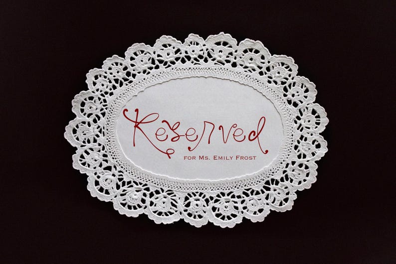 May include: White, oval-shaped doily with a delicate lace pattern. The center features the word "Reserved" in red script, followed by "for Ms. Emily Frost". The doily is set against a dark brown background.