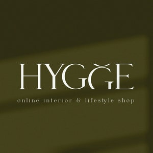K&ouml;nnte beinhalten: Wei&szlig;er Text auf gr&uuml;nem Hintergrund lautet "HYGGE" mit einer Tilde &uuml;ber dem "G". Unter dem Text steht "Online-Shop f&uuml;r Innenarchitektur & Lifestyle". Der Text wurde von "Cypria Studio" entworfen.