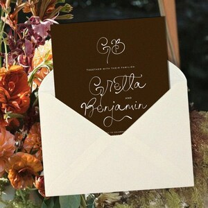May include: A wedding invitation card in a cream envelope. The dark brown card features white calligraphy with the names Gretta and Benjamin. The invitation is set against a backdrop of colorful flowers, including orange dahlias.