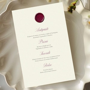 May include: A cream-colored menu card with a burgundy wax seal, featuring Italian text. The menu is placed on a white, scalloped-edge plate, with floral accents. The menu lists antipasti, primi, secondi, and dolce.