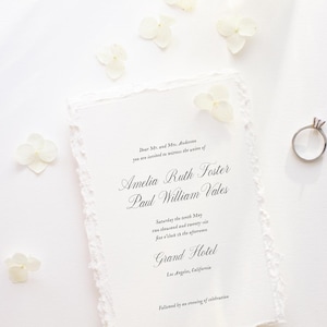 May include: A wedding invitation with torn edges, featuring the names Amelia Ruth Foster and Paul William Vales, along with a diamond ring and white flowers. The invitation is on a white surface, with elegant script.
