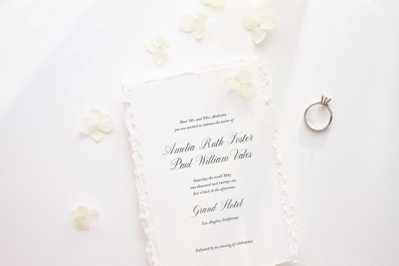 May include: A wedding invitation with torn edges, featuring the names Amelia Ruth Foster and Paul William Vales, along with a diamond ring and white flowers. The invitation is on a white surface, with elegant script.