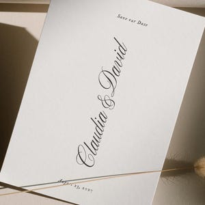 May include: A white invitation card with elegant script that reads "Claudia & David" and "Save our Date". The date "August 23, 2027" is also printed on the card. A dried flower stem is in the foreground.