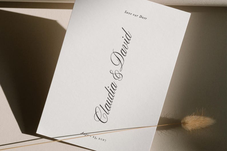 May include: A white invitation card with elegant script that reads "Claudia & David" and "Save our Date". The date "August 23, 2027" is also printed on the card. A dried flower stem is in the foreground.