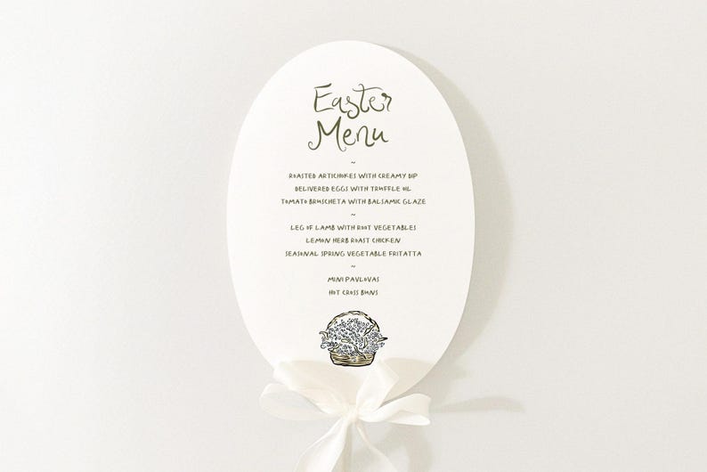 May include: White oval card with a handwritten Easter menu. The menu includes roasted artichokes,  believeres eggs,  tomato brichetta, leg of lamb, lemon herb roast chicken, seasonal spring vegetable frittata, mini pavlova, and hot cross buns. A small illustration of a basket of flowers is at the bottom of the card.