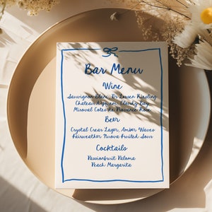 May include: A white bar menu with blue handwritten text. The menu lists wine, beer, and cocktails. The text includes: Sauvignon Blanc, Dr. Loosen Riesling, Chateau d'yquem, Cloudy Bay, Miroval Cotes de Provence Rose, Crystal Clear Lager, Amber Waves, Fairweather Nuova Fruited Sour, Passionfruit Paloma, and Peach Margarita.