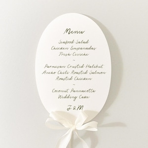 Puede incluir: Una tarjeta de men&uacute; de boda ovalada con fondo blanco y escritura elegante. El men&uacute; enumera platos como ensalada de mariscos y pollo asado. Una cinta blanca est&aacute; atada en la parte inferior, y las iniciales "J&M" est&aacute;n en la parte inferior.