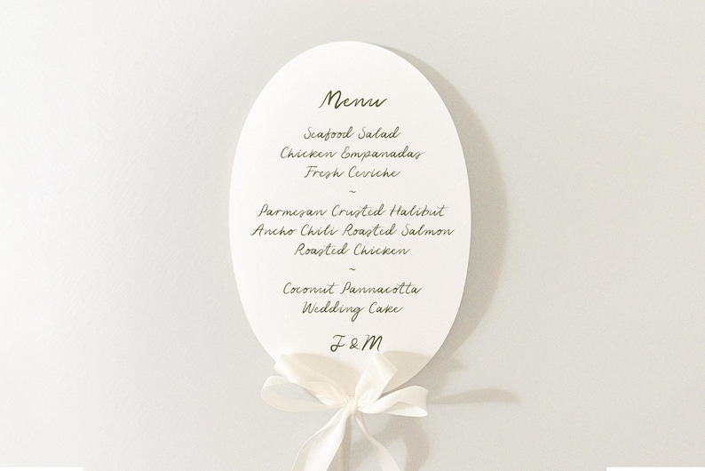Puede incluir: Una tarjeta de men&uacute; de boda ovalada con fondo blanco y escritura elegante. El men&uacute; enumera platos como ensalada de mariscos y pollo asado. Una cinta blanca est&aacute; atada en la parte inferior, y las iniciales "J&M" est&aacute;n en la parte inferior.