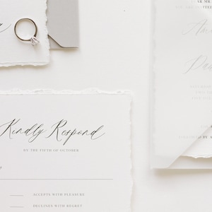 May include: A white RSVP card with a handwritten message that reads "Kindly Respond by the Fifth of October". The card has two options for guests to choose from: "Accepts with Pleasure" and "Declines with Regret".