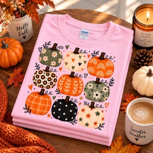 Peut inclure: Un t-shirt rose clair avec une impression graphique de neuf citrouilles aux motifs variés, incluant des fleurs, des taches de léopard, des pois, des cœurs et des arcs-en-ciel, agrémenté de petits cœurs, d'étoiles et de feuilles éparpillées.