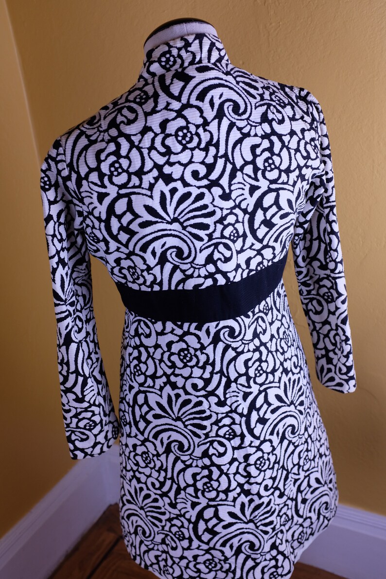 K&ouml;nnte beinhalten: Ein schwarz-wei&szlig;es Kleid mit Blumenmuster. Das Kleid hat lange &Auml;rmel und einen taillierten Oberteil. Das Kleid ist aus einem weichen, dehnbaren Stoff.
