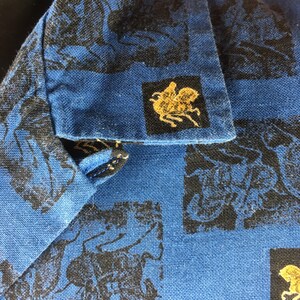 May include: Blue cotton shirt with a black and gold print of a repeated design. The design features a horse and rider.