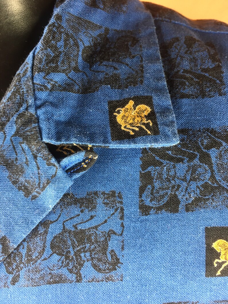 May include: Blue cotton shirt with a black and gold print of a repeated design. The design features a horse and rider.