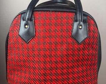 May include: A vintage handbag with a red and brown woven pattern. The bag features black leather trim, handles, and accents. The handles are attached with gold-colored rivets. The bag is sitting on a brown surface.