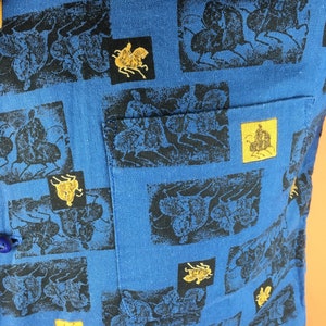 May include: A blue cotton shirt with a repeating pattern of black silhouettes of people riding horses. The pattern is framed in a rectangular shape and has a gold outline.