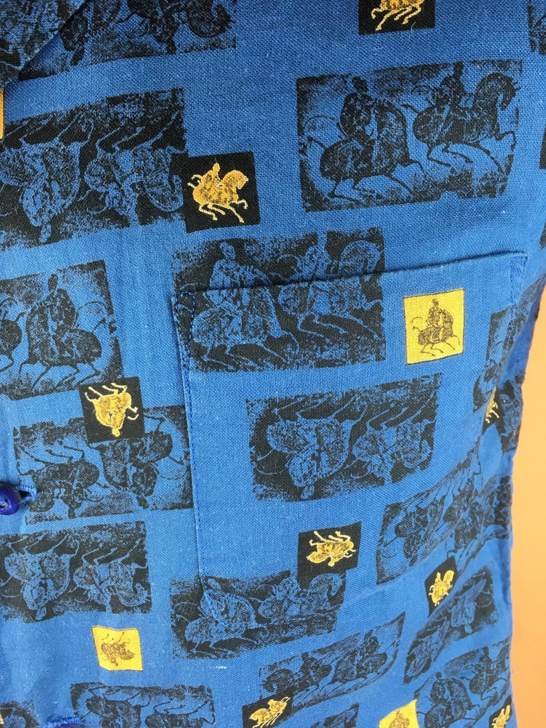 May include: A blue cotton shirt with a repeating pattern of black silhouettes of people riding horses. The pattern is framed in a rectangular shape and has a gold outline.