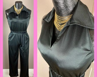Puede incluir: Mono negro con cuello y cintura ceñida. El mono sin mangas tiene un diseño de pierna ancha y un cinturón a juego. Un collar dorado está drapeado alrededor del cuello del maniquí. El mono se exhibe en un maniquí sobre un fondo rosa.