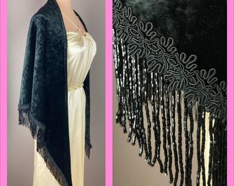 May include: A dark green velvet shawl with fringe and beaded trim. The shawl is draped over a cream-colored dress. The fringe and trim are black. The shawl is a vintage style.