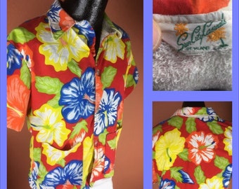 Op de afbeelding: Een overhemd met korte mouwen en knopen met een levendig bloemenpatroon. Het overhemd heeft grote blauwe, gele en witte bloemen op een rode achtergrond, met groene bladeren. Het label luidt "Sun City Casuals".