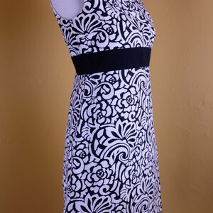 K&ouml;nnte beinhalten: Ein &auml;rmelloses, schwarz-wei&szlig;es Kleid mit Blumenmuster. Das Kleid hat einen schwarzen G&uuml;rtel und eine figurbetonte Silhouette.
