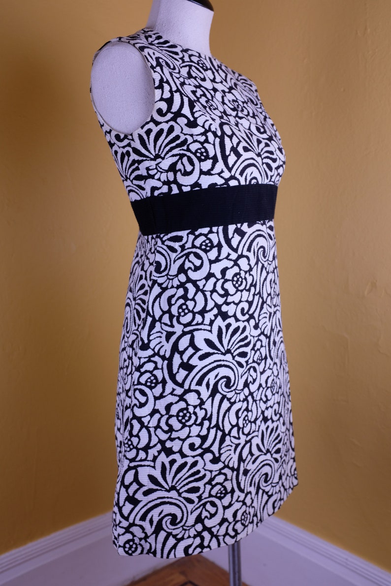 K&ouml;nnte beinhalten: Ein &auml;rmelloses, schwarz-wei&szlig;es Kleid mit Blumenmuster. Das Kleid hat einen schwarzen G&uuml;rtel und eine figurbetonte Silhouette.