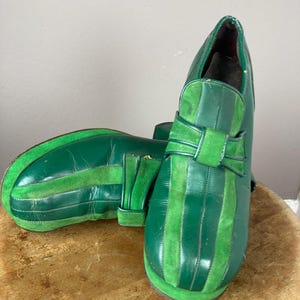 May include: A pair of green platform shoes with vertical stripes and a bow detail. The shoes have a rounded toe and a thick sole. The design incorporates both smooth and textured green materials.