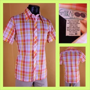 Puede incluir: Una camisa de manga corta a cuadros con cuello abotonado. La camisa es de cuadros rosa, naranja y blanco. La camisa tiene una etiqueta que dice "Men's Hukap Huk-A-Fit 65% Polyester 35% Cotton Machine Wash Tumble Dry Low Setting Remove Immediately Do Not Use Bleach Made in Hong Kong RN 34104 Size: M".