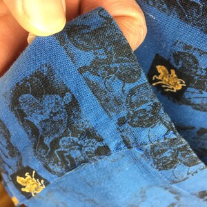 May include: Close-up of a blue fabric with a black and gold pattern. The fabric has a repeating pattern of a black silhouette of a person riding a horse. The pattern is surrounded by a gold border.