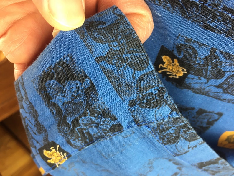 May include: Close-up of a blue fabric with a black and gold pattern. The fabric has a repeating pattern of a black silhouette of a person riding a horse. The pattern is surrounded by a gold border.