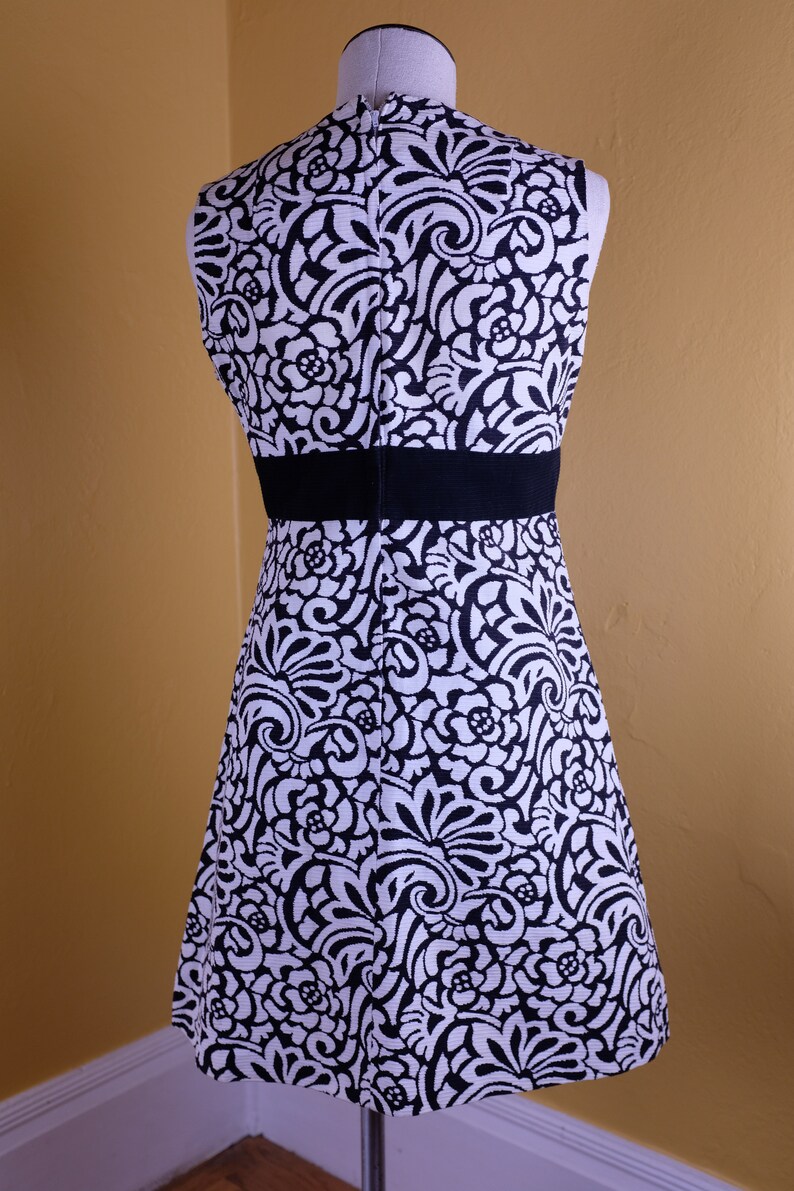 K&ouml;nnte beinhalten: Ein &auml;rmelloses schwarz-wei&szlig;es Kleid mit Blumenmuster. Das Kleid hat einen schwarzen Tailleng&uuml;rtel und ein figurbetontes Oberteil.