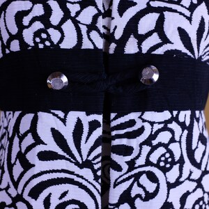 K&ouml;nnte beinhalten: Schwarze Jacke mit wei&szlig;em Blumenmuster, einem schwarzen Band und silbernen Kn&ouml;pfen.