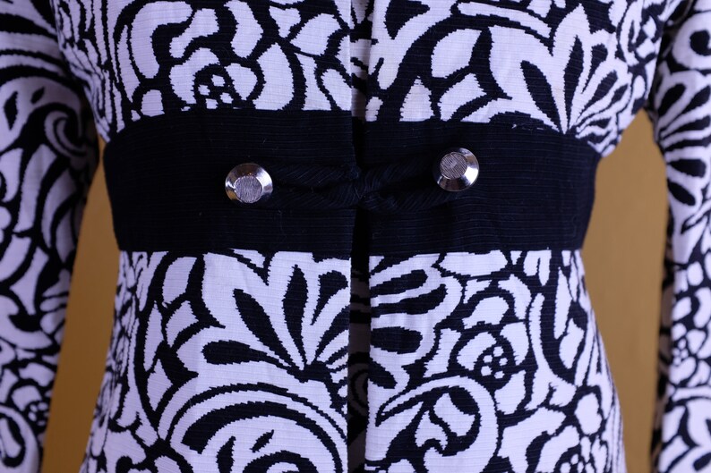K&ouml;nnte beinhalten: Schwarze Jacke mit wei&szlig;em Blumenmuster, einem schwarzen Band und silbernen Kn&ouml;pfen.