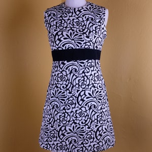 K&ouml;nnte beinhalten: Ein &auml;rmelloses schwarz-wei&szlig;es Kleid mit floralem Muster und einem schwarzen G&uuml;rtel. Das Kleid befindet sich auf einer Schaufensterpuppe vor einer gelben Wand.