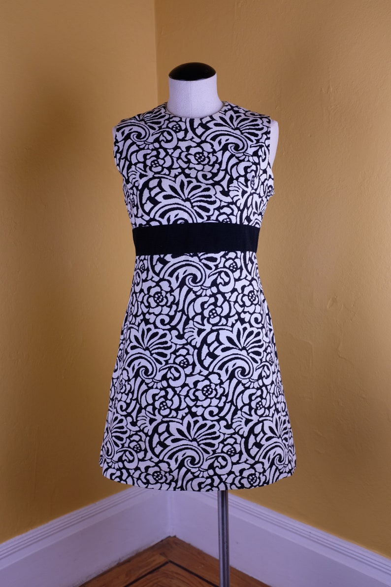 K&ouml;nnte beinhalten: Ein &auml;rmelloses schwarz-wei&szlig;es Kleid mit floralem Muster und einem schwarzen G&uuml;rtel. Das Kleid befindet sich auf einer Schaufensterpuppe vor einer gelben Wand.