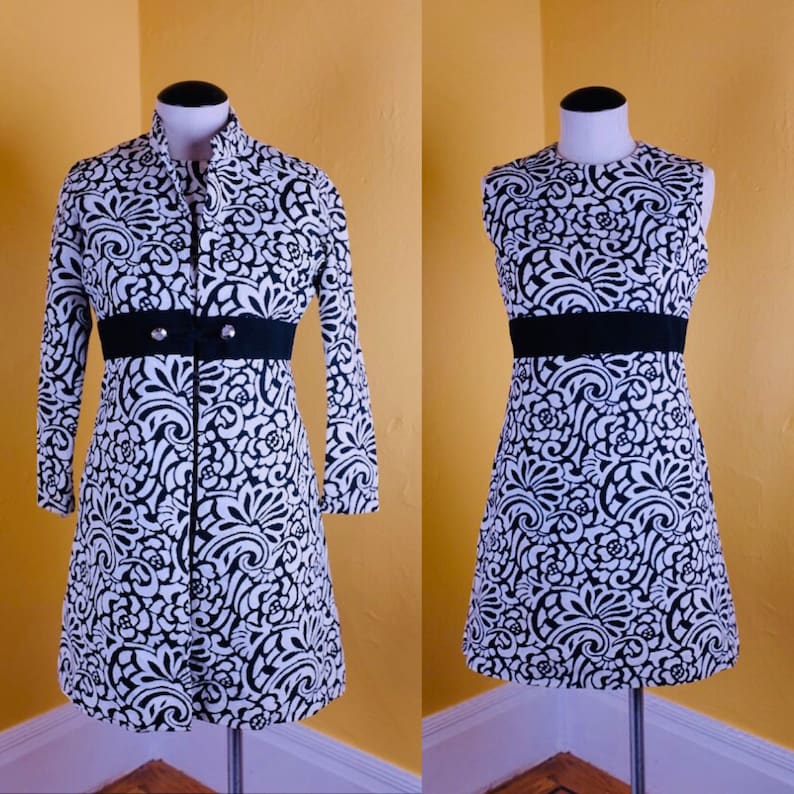 K&ouml;nnte beinhalten: Ein schwarz-wei&szlig; gemustertes Kleid mit einem schwarzen G&uuml;rtel und einer passenden Jacke. Das Kleid und die Jacke haben ein florales Design.