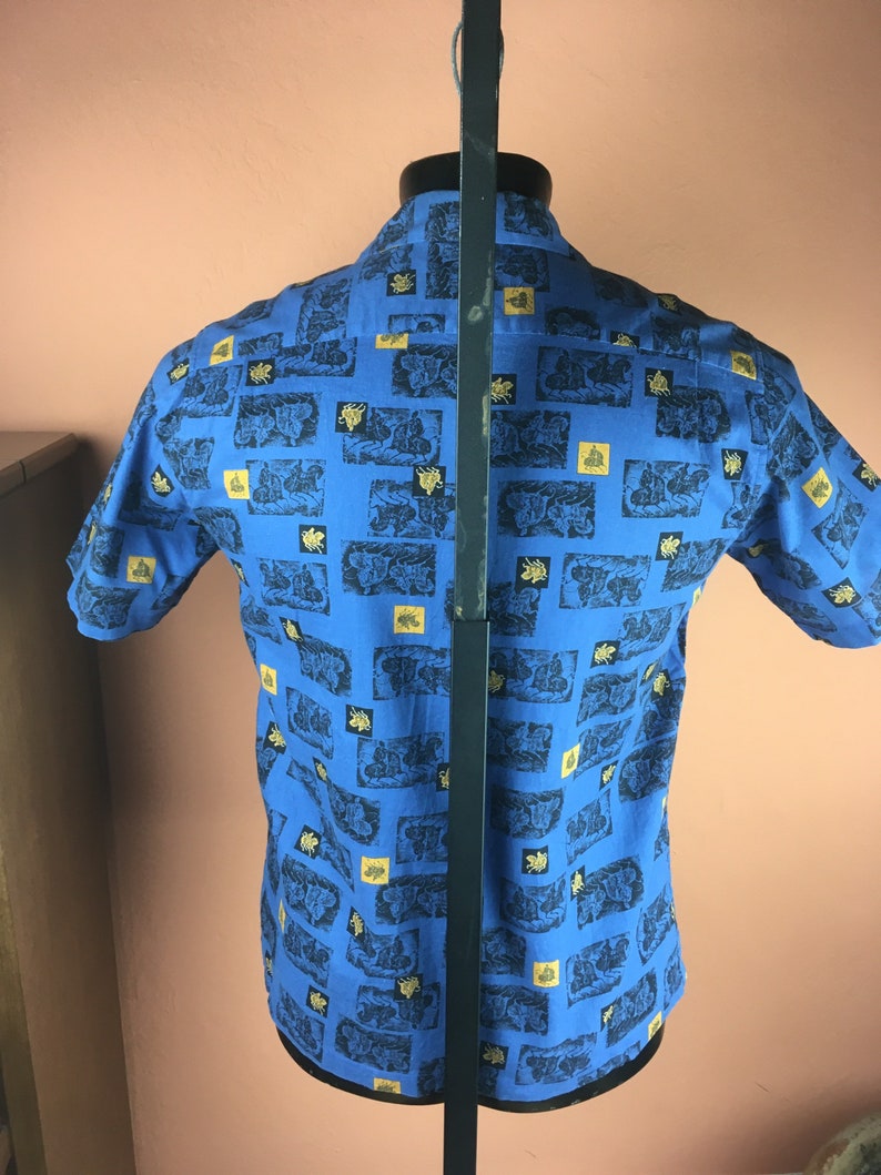 May include: A blue short-sleeved shirt with a repeating pattern of black and gold images. The shirt is on a mannequin.
