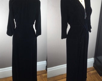 May include: A black velvet dress with a V-neckline and a knot detail at the waist. The dress has long sleeves and a floor-length skirt.