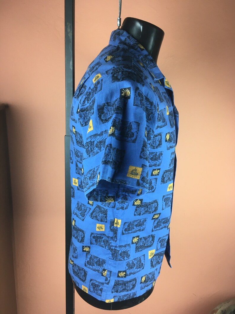 May include: A blue short-sleeved button-down shirt with a gold and black patterned design.