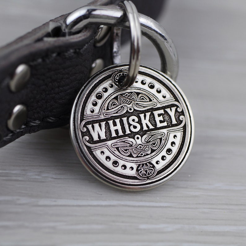 May include: A silver dog tag with an ornate design and the word "WHISKEY" engraved in black lettering.