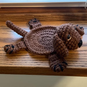 May include: A brown crocheted dog-shaped coaster with black eyes and nose. The coaster is lying on a wooden surface.