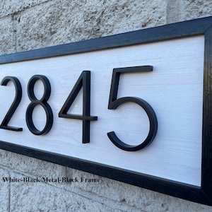 Könnte beinhalten: Ein weißes Holzschild mit schwarzem Rahmen und schwarzen Metallziffern, die "2845" anzeigen.