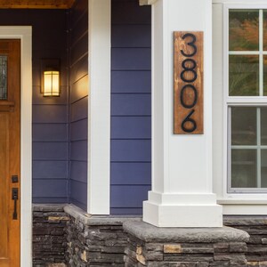 May include: A wooden house number sign with black metal numbers. The sign is mounted on a white column and reads "3806".