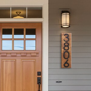 May include: A wooden house number sign with black metal numbers. The sign reads "3806" and is mounted on a gray siding wall.