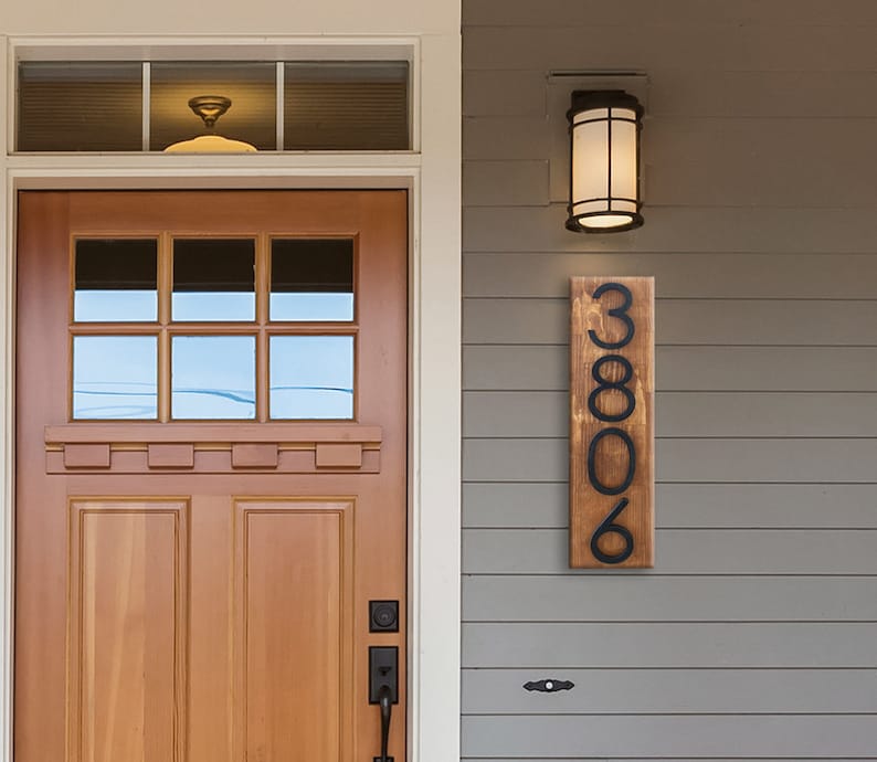 May include: A wooden house number sign with black metal numbers. The sign reads "3806" and is mounted on a gray siding wall.
