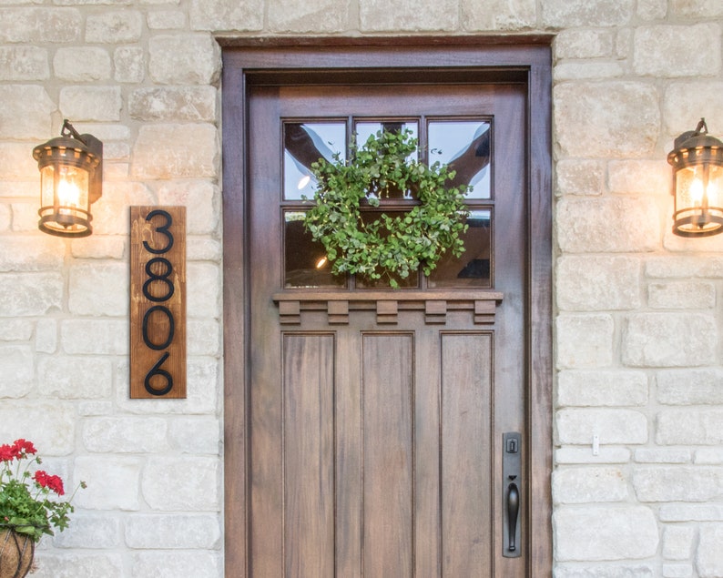 May include: A wooden house number sign with black metal numbers reading "3806" mounted on a white brick wall. A wooden door with a glass window pane and a wreath is to the right of the sign.